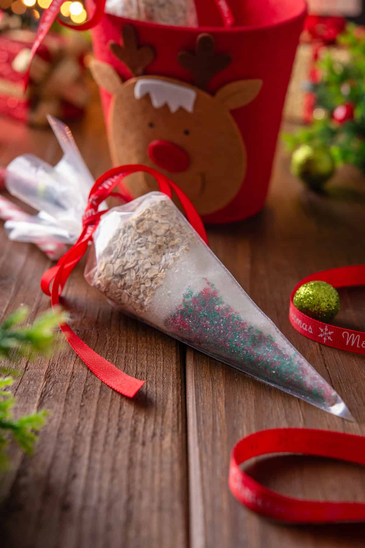 A piping bag filled with reindeer food tied with a red bow, next to candy canes and Christmas ornaments on a wooden tabletop.