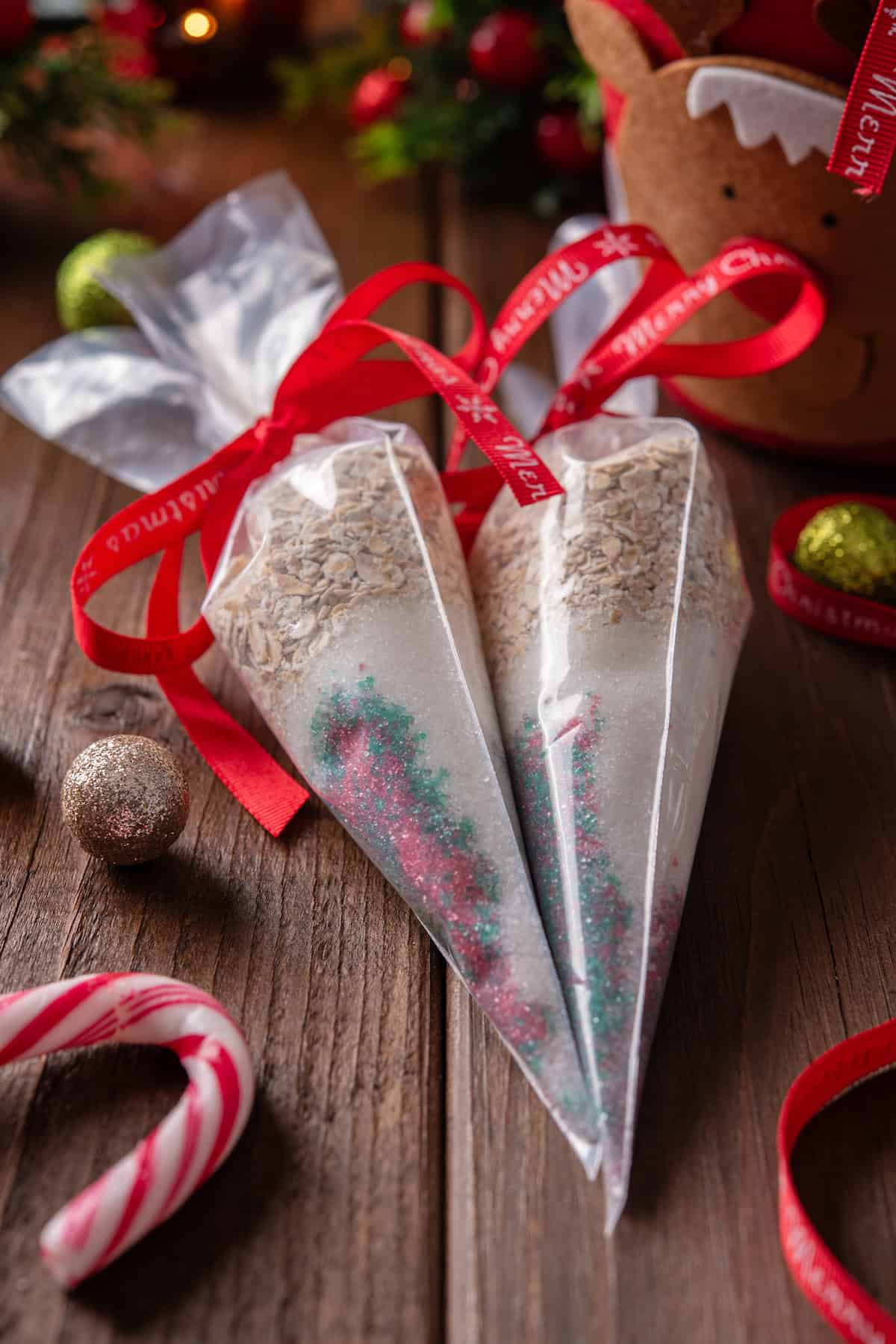 Two side by side piping bags filled with reindeer food tied with red bows, next to Christmas ornaments on a wooden tabletop.