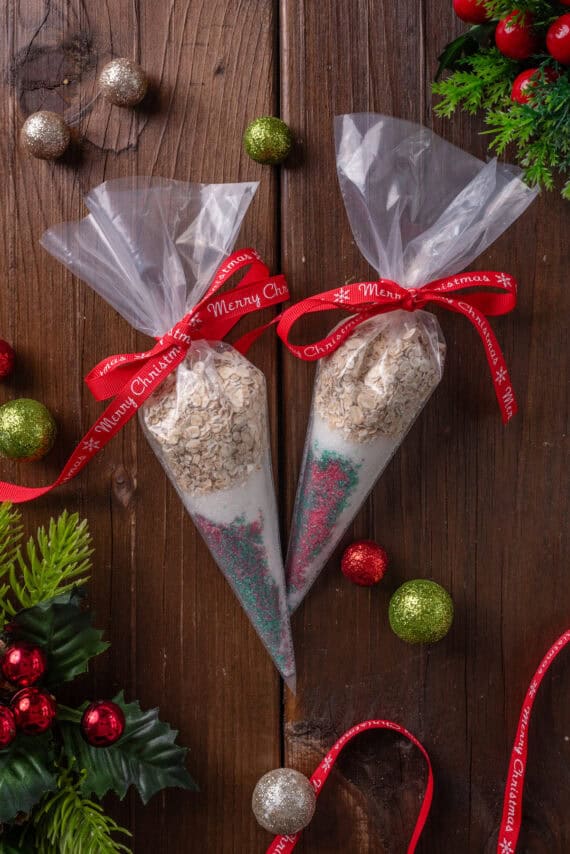 Overhead view of two piping bags filled with reindeer food tied with red bows, surrounded by scattered Christmas ornaments on a wooden tabletop.