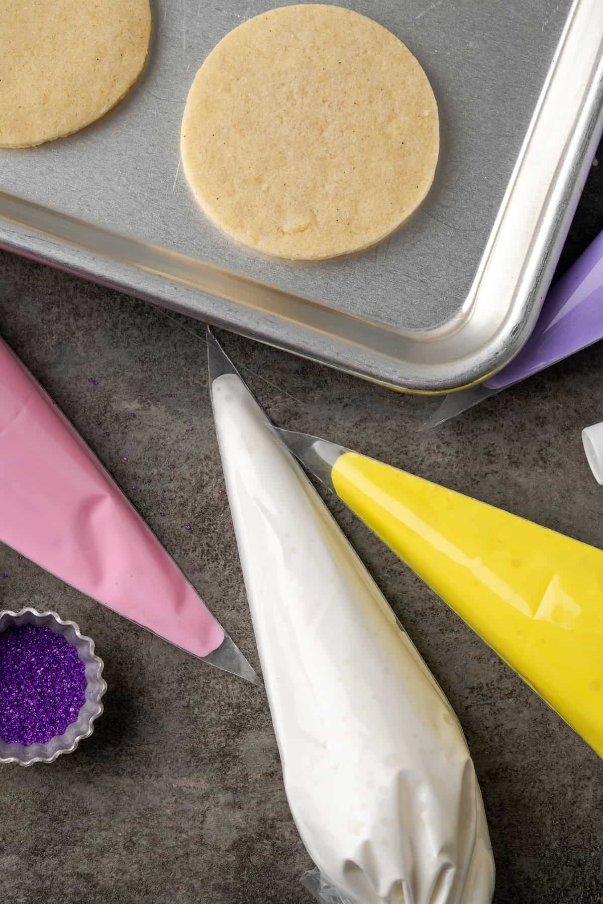 Overhead view of assorted piping bags filled with pink, white, and yellow royal icing on a grey countertop, next to round sugar cookies on a baking sheet.