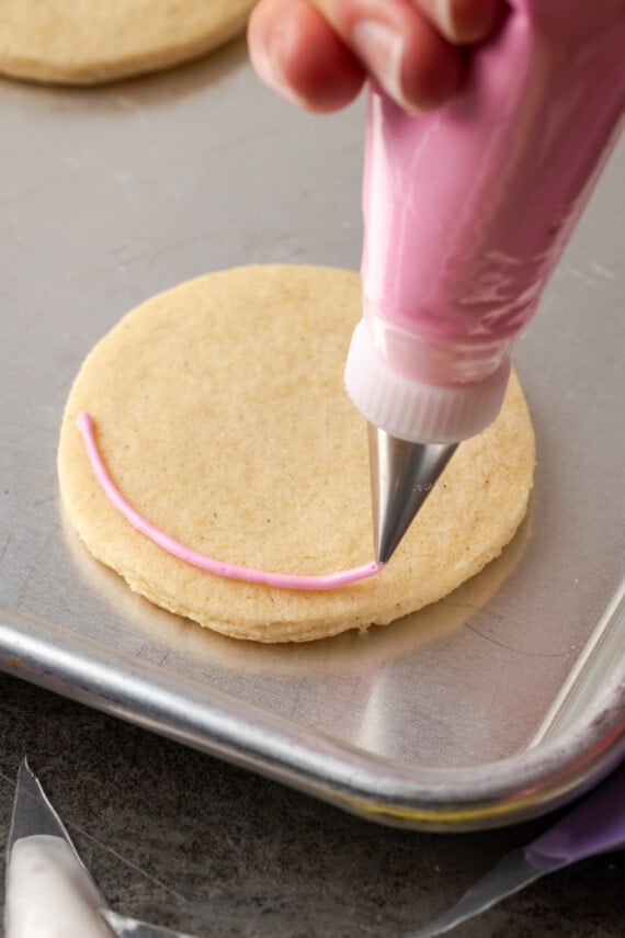 A piping tip beginning to pipe an outline of pink royal icing around the outside edge of a round sugar cookie on a baking sheet.