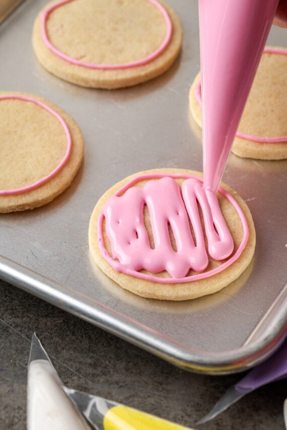 A piping tip flooding the outline on a round sugar cookie with pink royal icing.