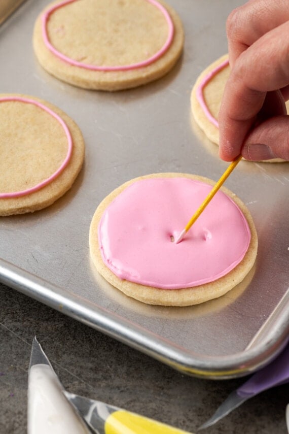 A hand using a toothpick to even out the surface of a sugar cookie flooded with pink royal icing.