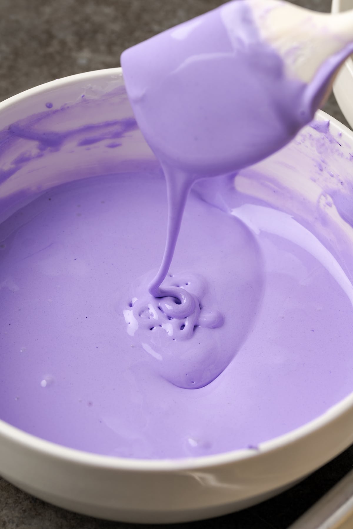 A rubber spatula drizzles purple royal icing into a bowl, showing the icing consistency needed for flooding.