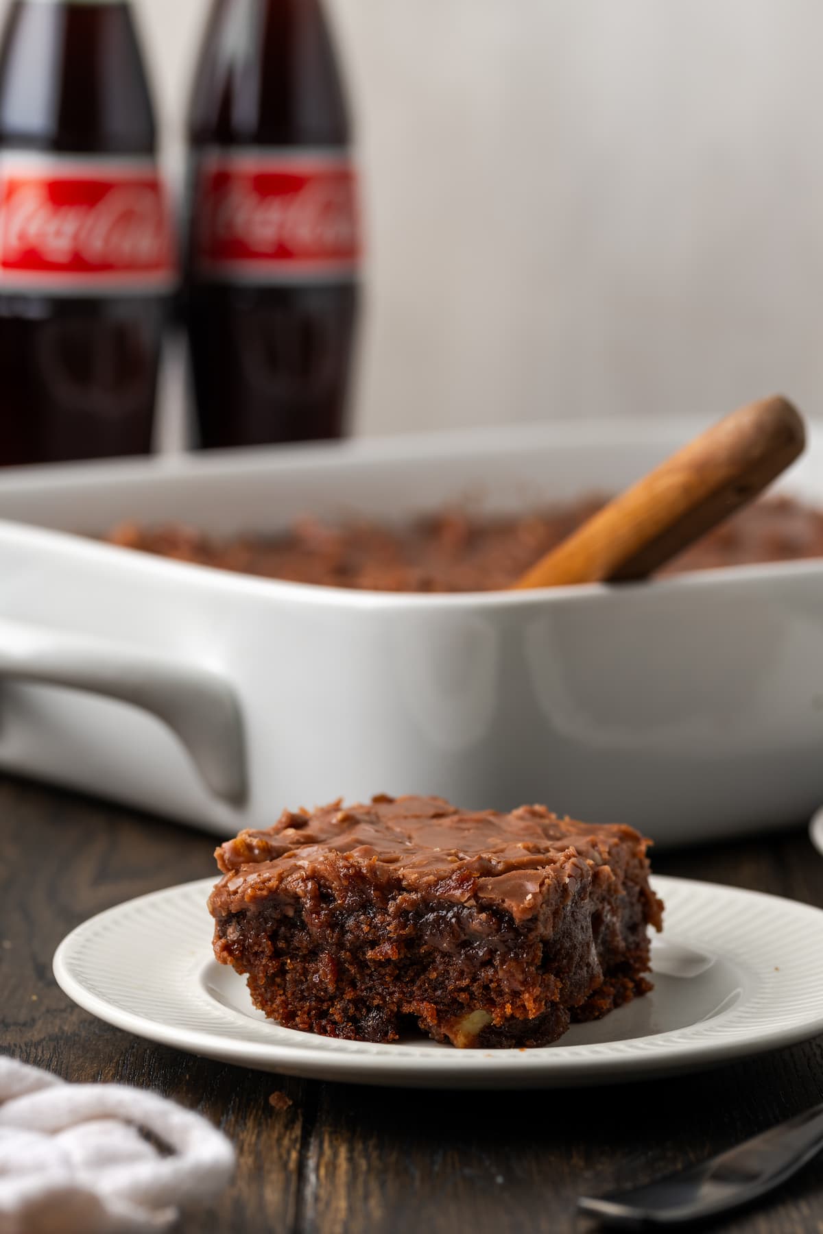 A slice of Coca Cola cake on a white plate, with the rest of the cake in a baking dish next to two Coke bottles in the background.