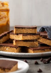 Side view of chocolate peanut butter bars stacked on a white plate, with a jar of peanut butter in the background.