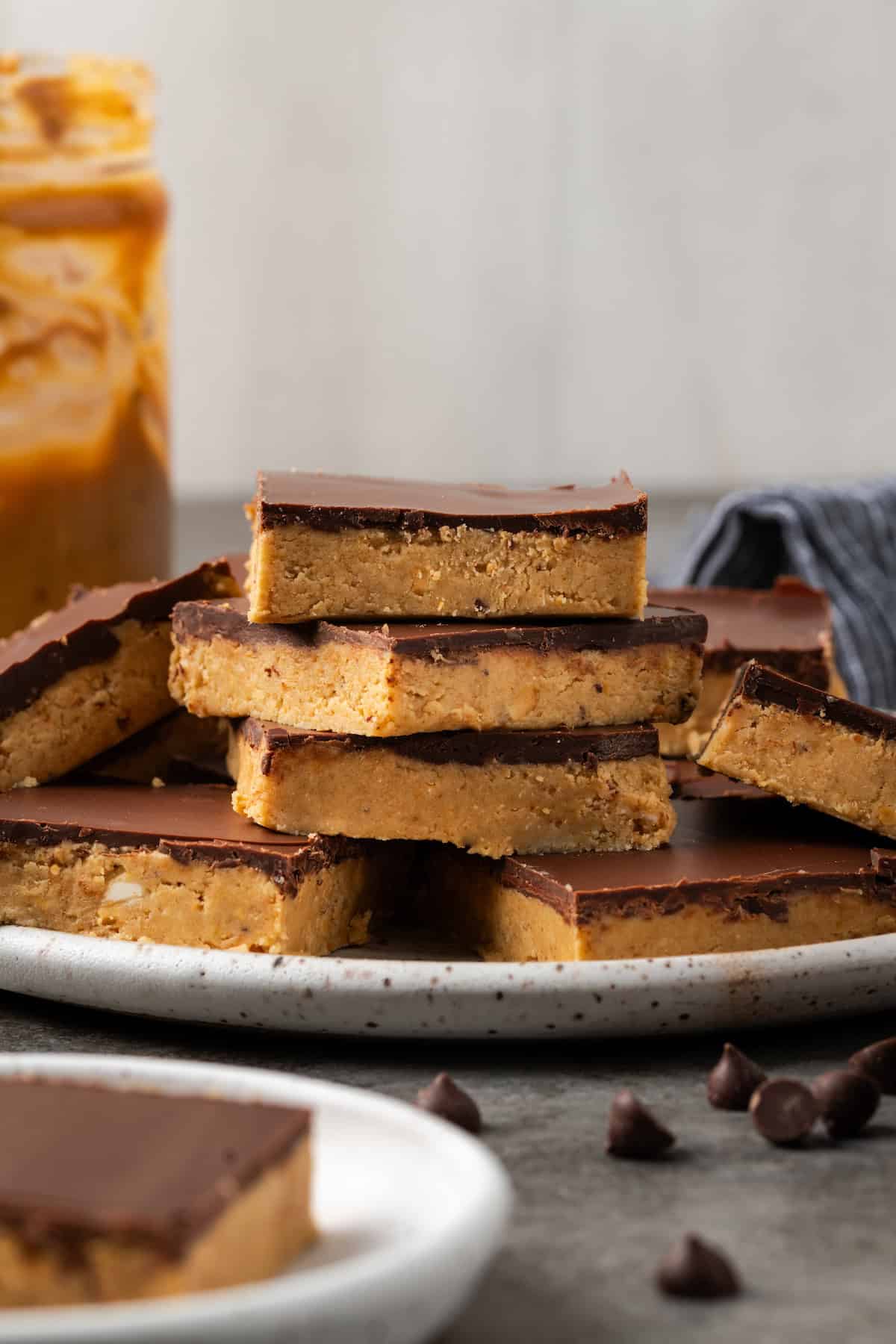 Side view of chocolate peanut butter bars stacked on a white plate, with a jar of peanut butter in the background.
