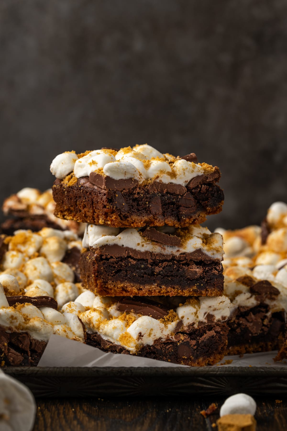 Three smores brownies stacked on a board lined with parchment paper, with more brownies in the background.