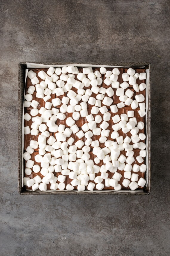 Mini marshmallows spread over chocolate bars layered on top of baked brownies in a square pan.