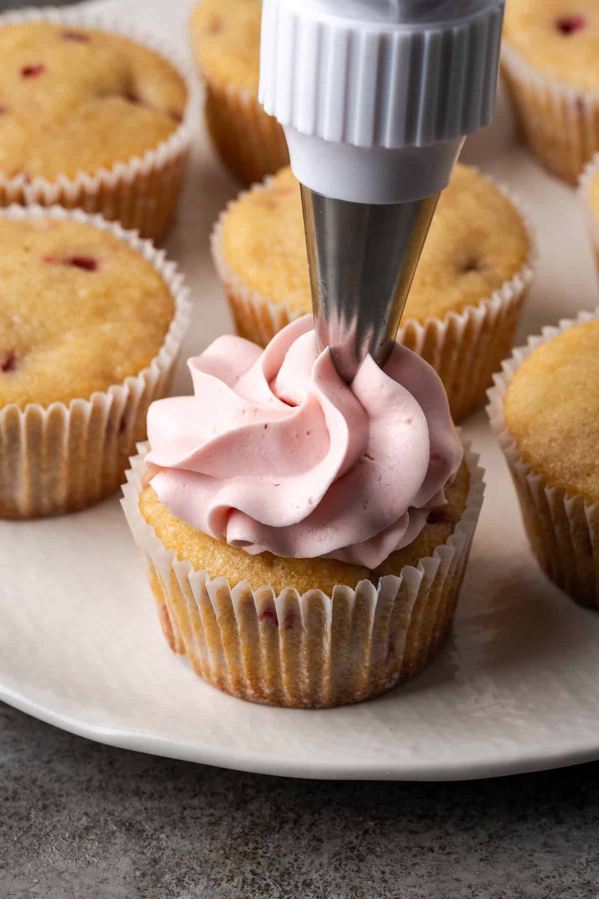 A piping tip piping a swirl of strawberry frosting onto a strawberry cupcake on a plate, with unfrosted cupcakes in the background.