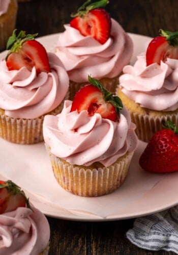 Four strawberry cupcakes frosted with swirls of strawberry Swiss meringue buttercream and topped with fresh strawberry halves on a plate.