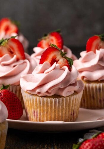 Side view of strawberry cupcakes frosted with swirls of strawberry Swiss meringue buttercream and topped with fresh strawberry halves on a plate.