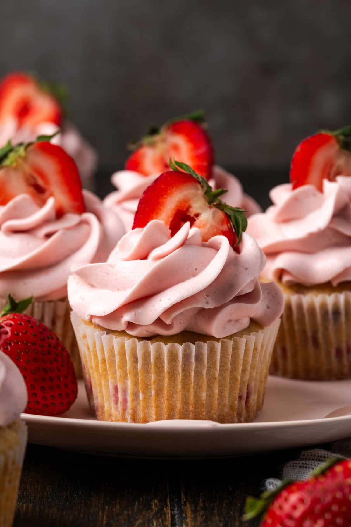 Side view of strawberry cupcakes frosted with swirls of strawberry Swiss meringue buttercream and topped with fresh strawberry halves on a plate.