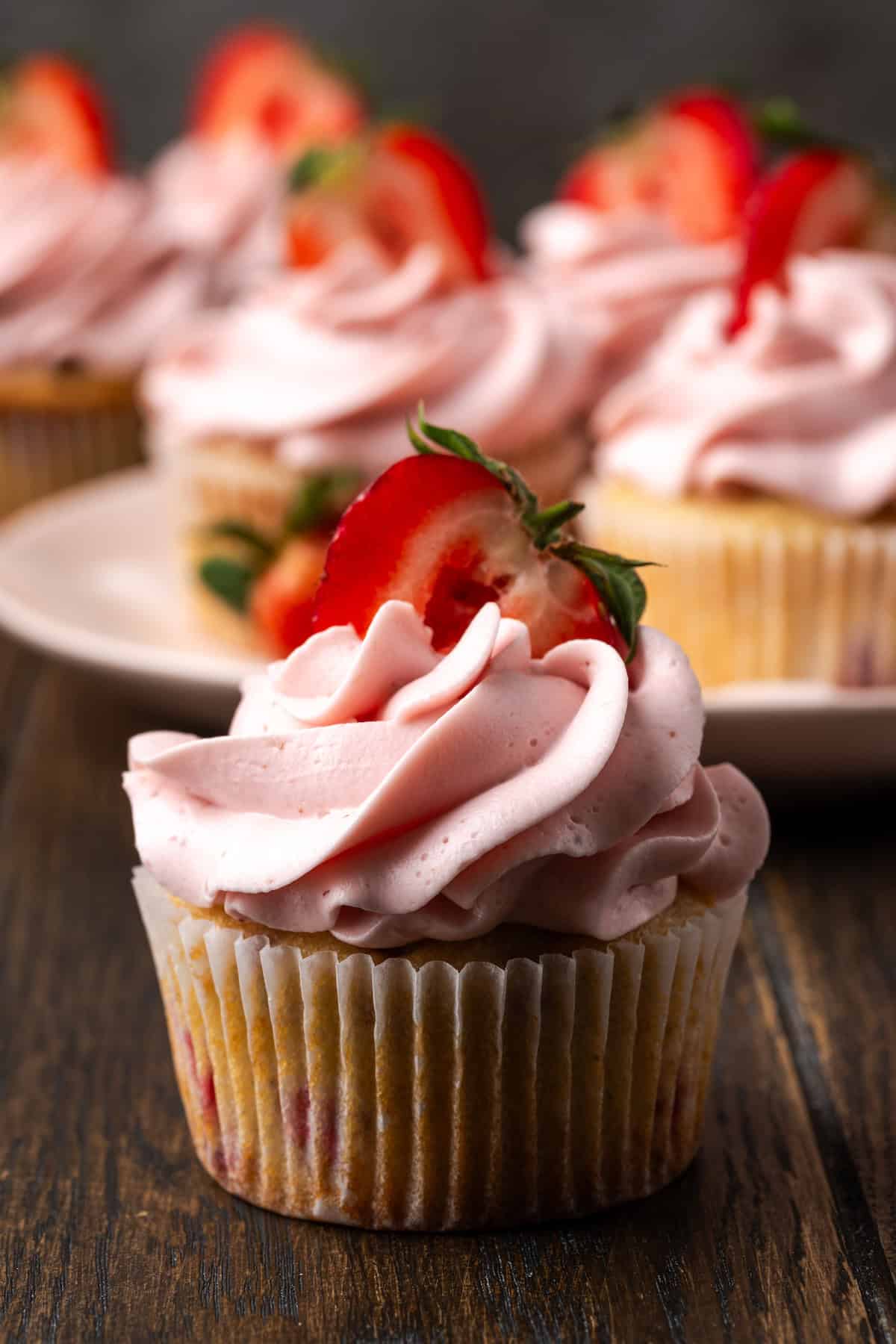 Close up of a strawberry cupcake frosted with strawberry Swiss meringue buttercream and topped with a fresh strawberry half, with more frosted cupcakes on a plate in the background.