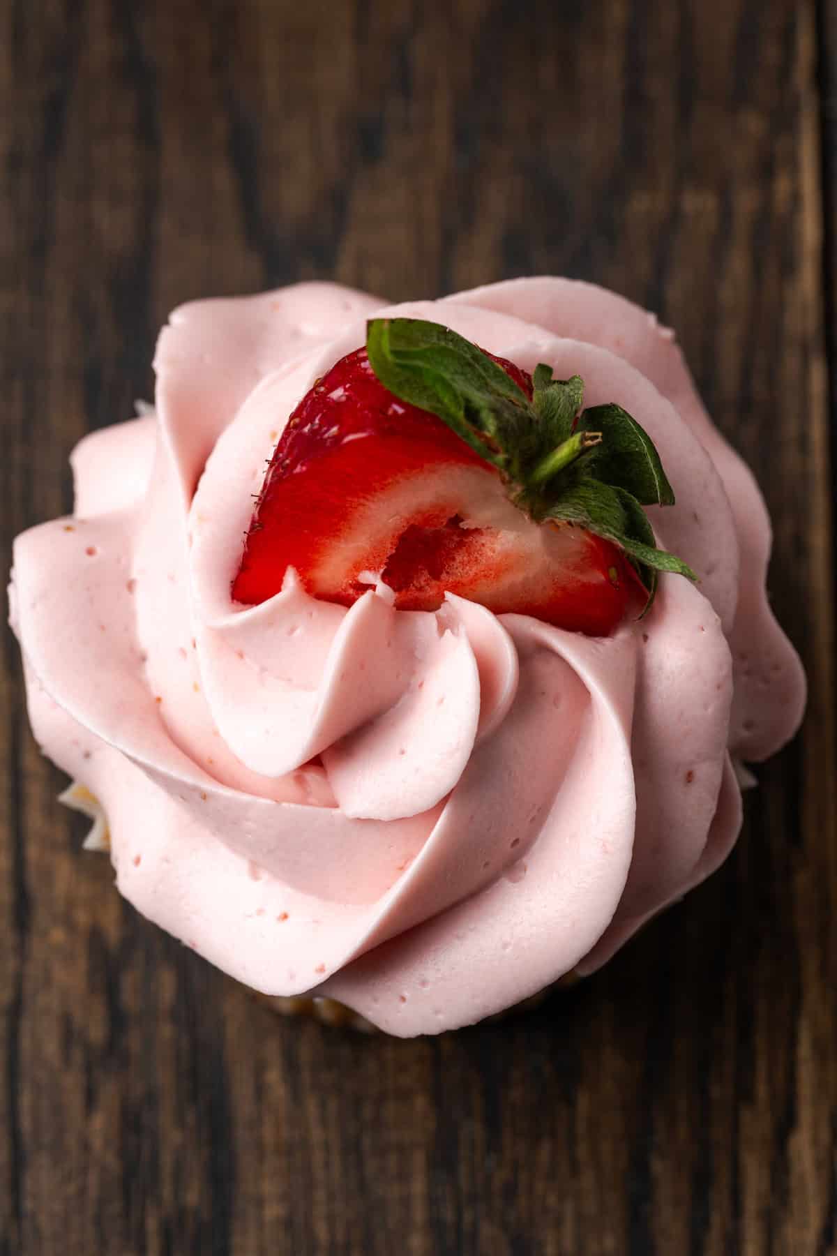 Overhead close up view of a frosted strawberry cupcake topped with a fresh strawberry half.