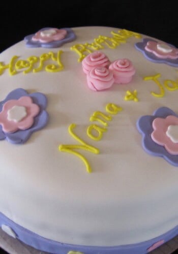Carrot Cake Decorated with White Fondant, Pink and Purple Flowers, and Yellow Happy Birthday Icing