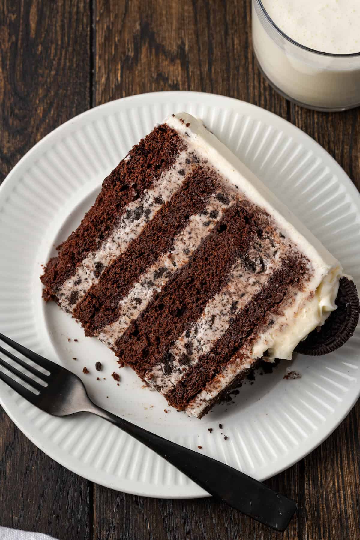 Overhead view of a slice of cookies and cream cake on a white plate next to a fork, beside a glass of milk.
