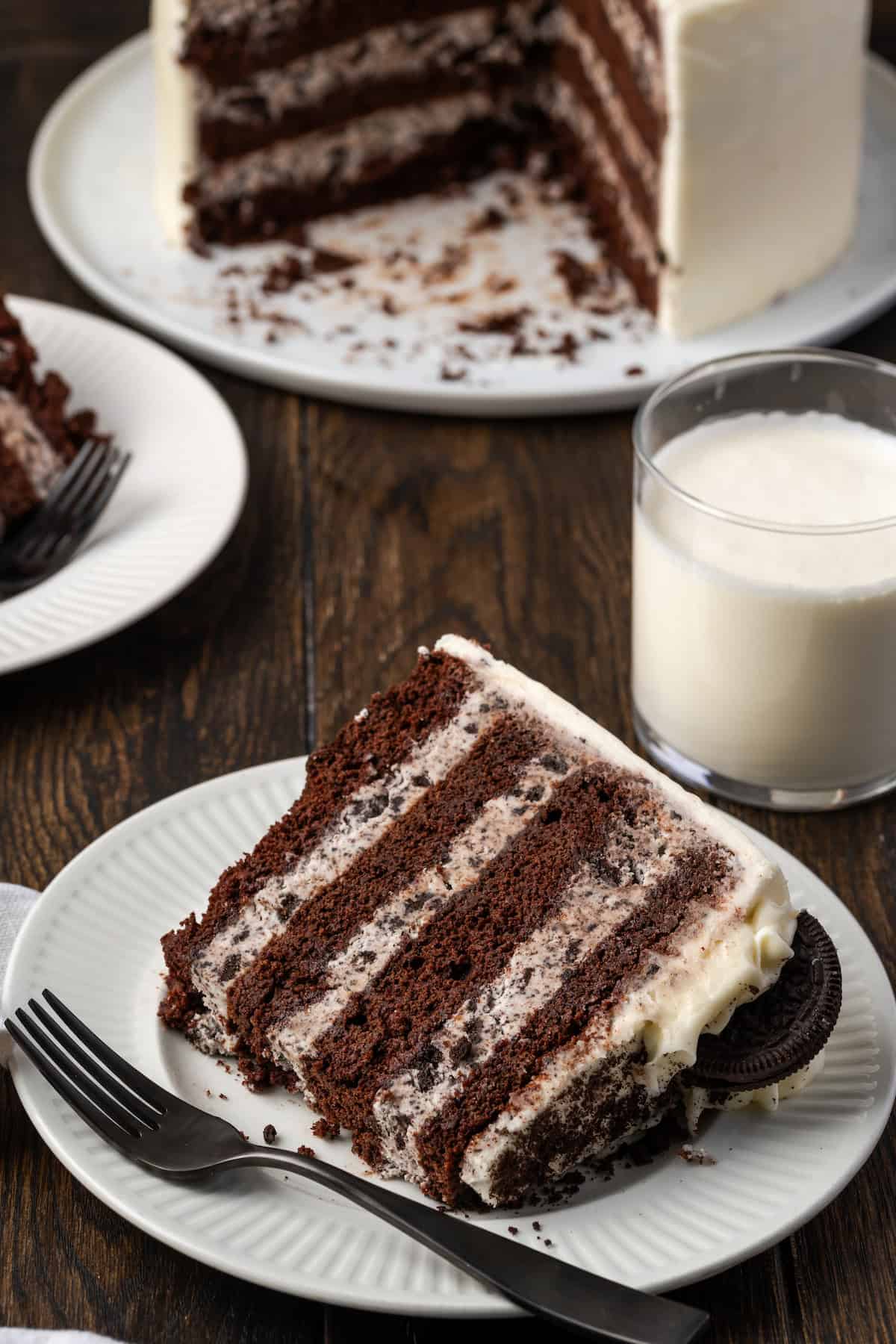 A slice of cookies and cream cake on a white plate next to a fork, with a glass of milk and the rest of the cake in the background.