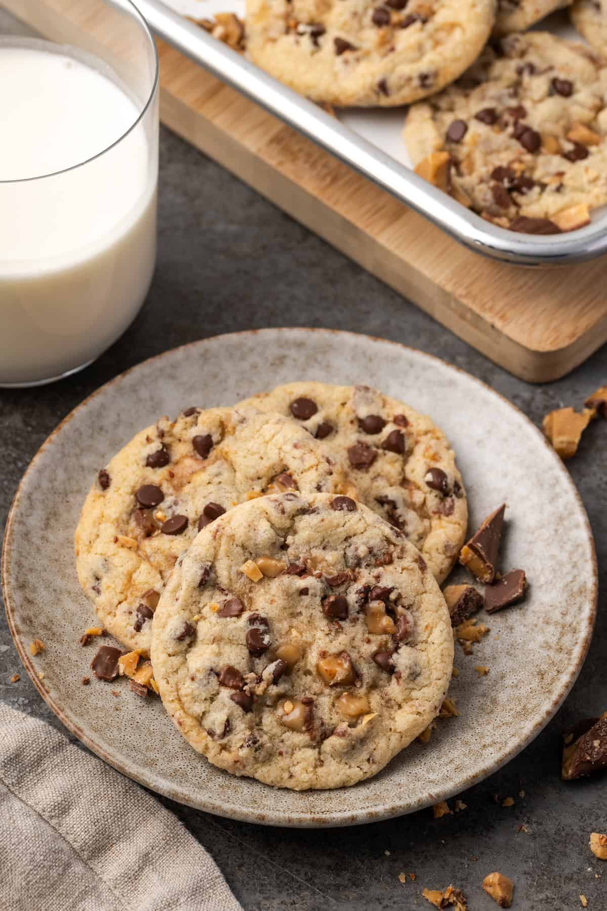 Toffee chocolate chip cookies stacked on a stoneware plate with a glass of milk in the background.