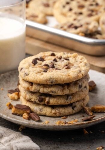Toffee chocolate chip cookies stacked on a stoneware plate with a glass of milk and a tray of cookies in the background.