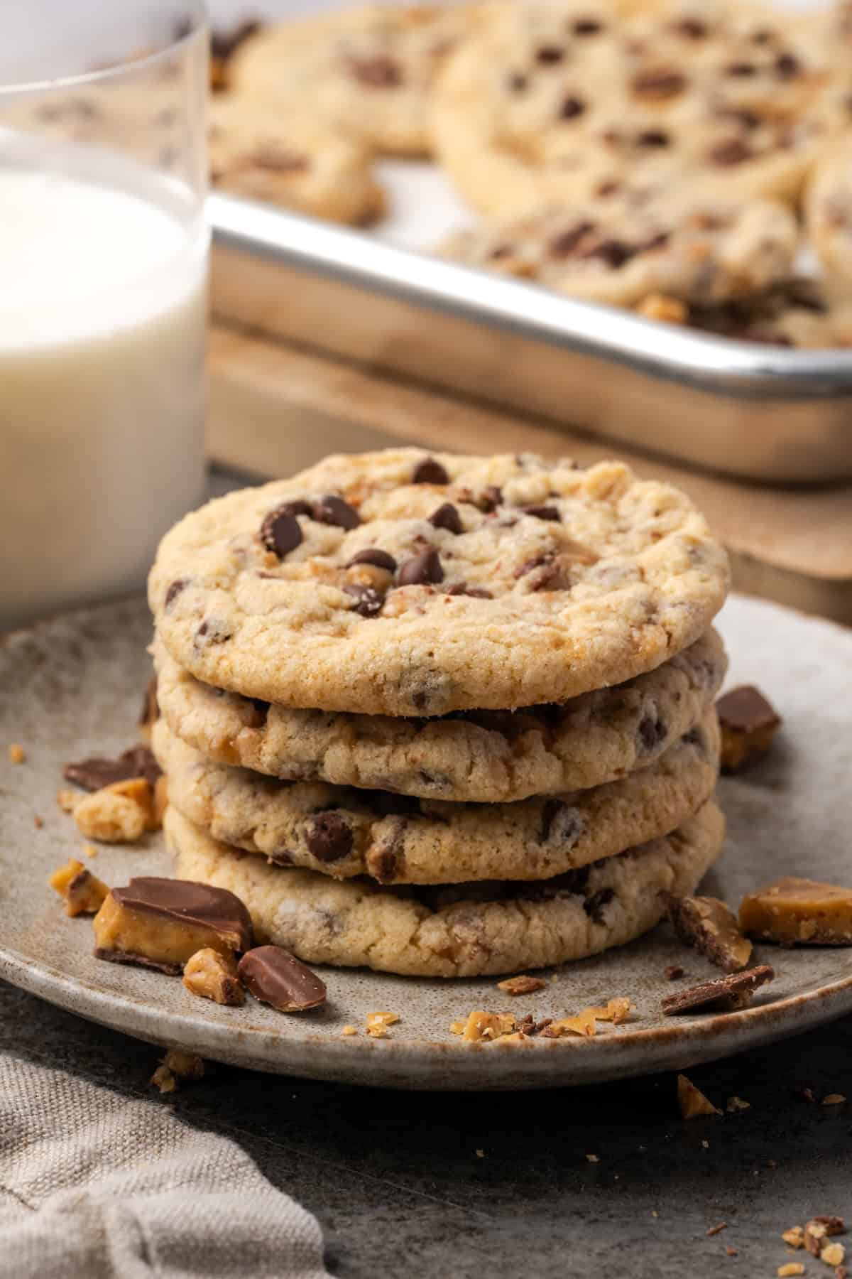 Toffee chocolate chip cookies stacked on a stoneware plate with a glass of milk and a tray of cookies in the background.