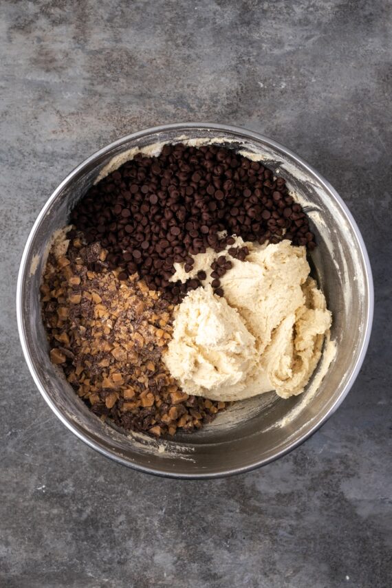 Toffee chips and chocolate chips added to cookie dough in a metal bowl.