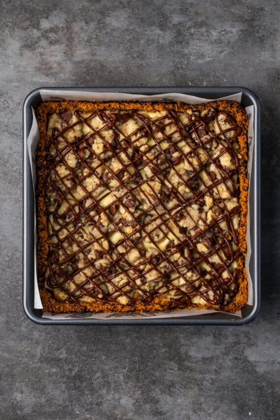 Baked chocolate chip cheesecake bars covered with drizzles of melted chocolate in a baking pan.