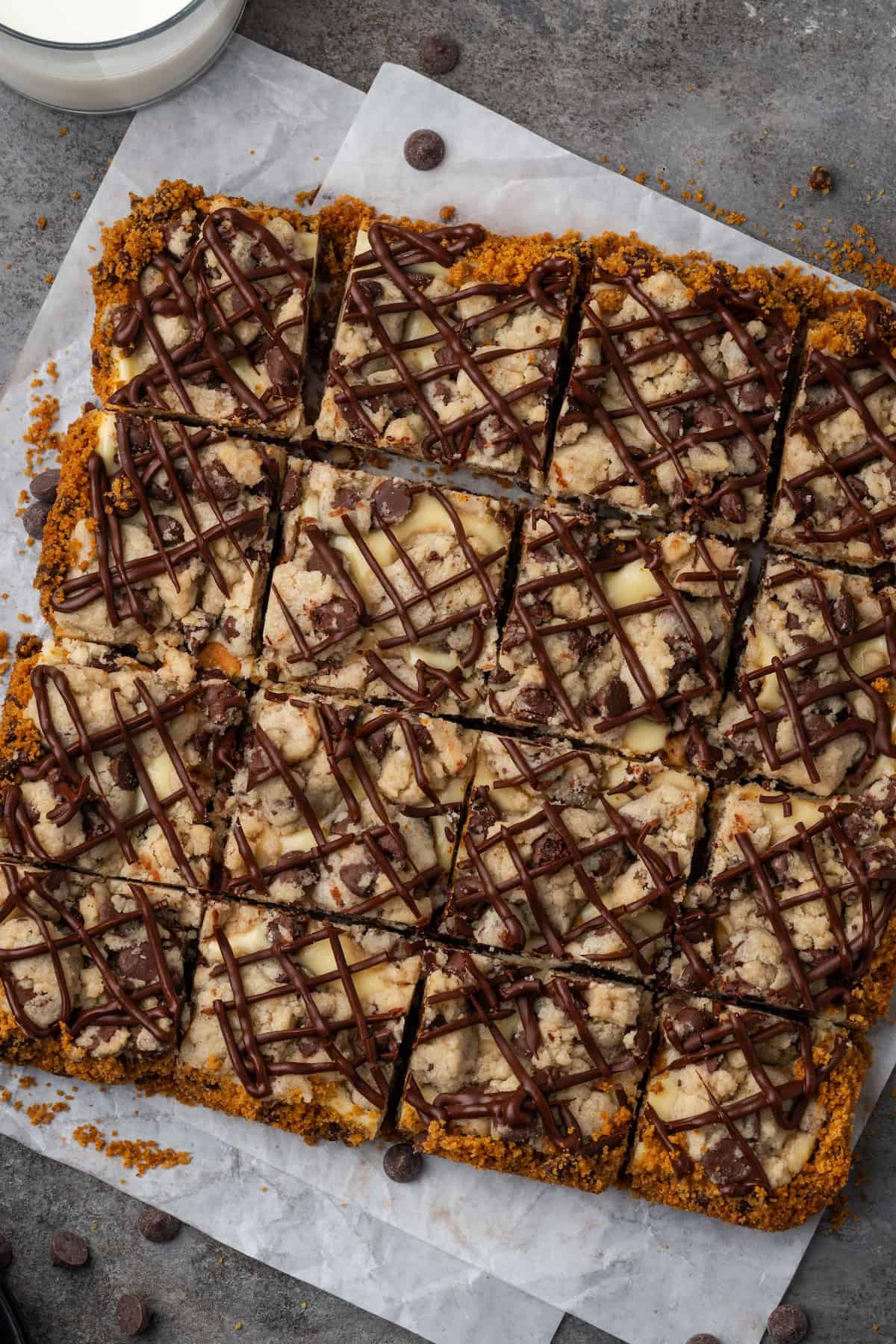 Overhead view of chocolate chip cheesecake bars covered with drizzles of melted chocolate on a sheet of parchment paper.
