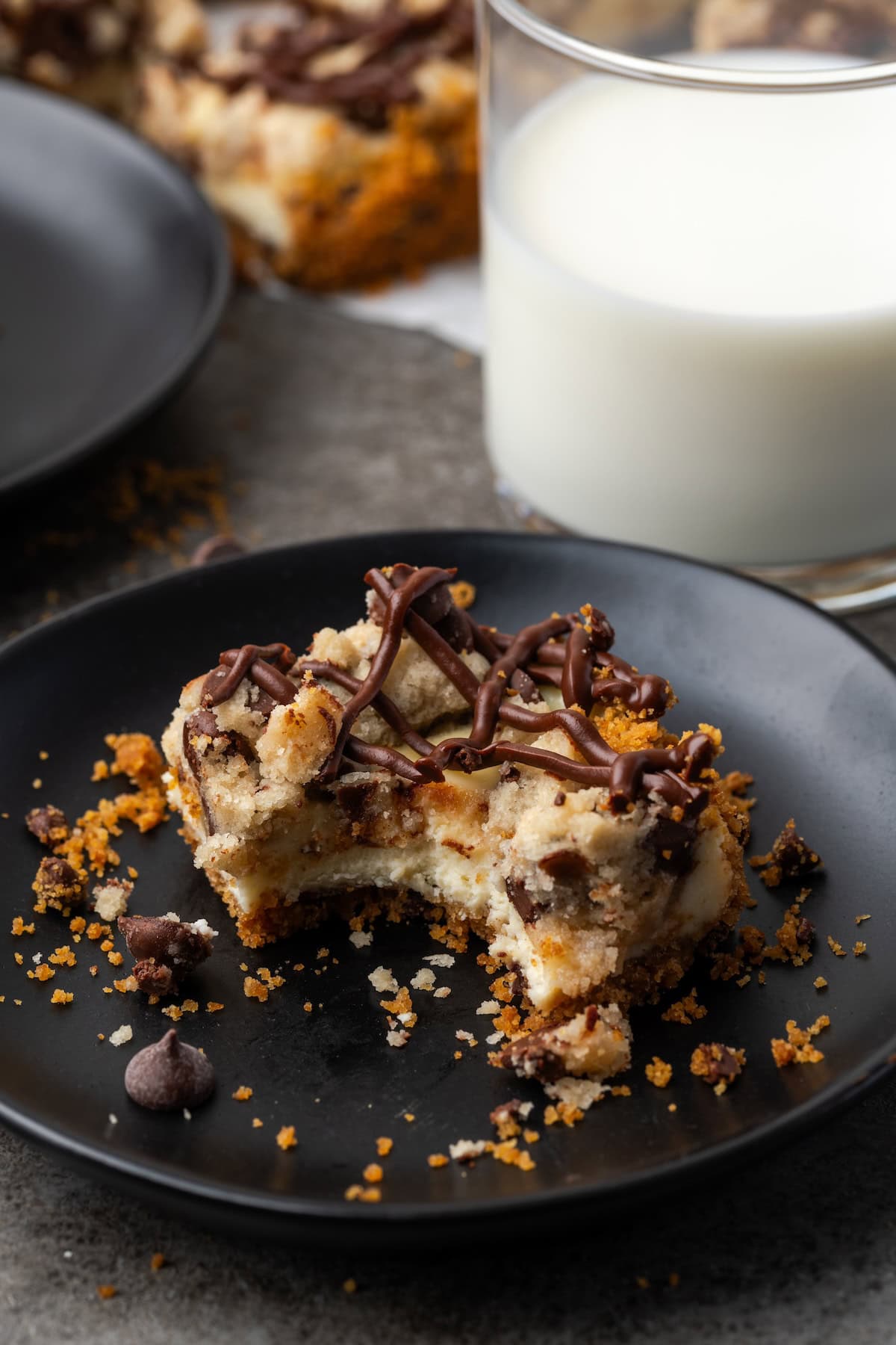 A chocolate chip cheesecake bar with a bite missing on a black stoneware plate, next to a glass of milk.