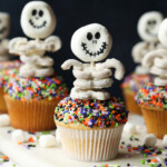 Multiple sprinkle covered cupcakes decorated with skeleton toppers made from white chocolate covered pretzels.