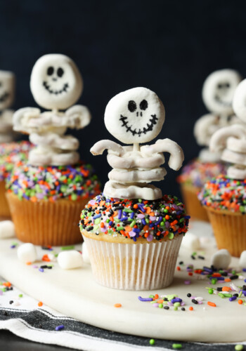 Multiple sprinkle covered cupcakes decorated with skeleton toppers made from white chocolate covered pretzels.
