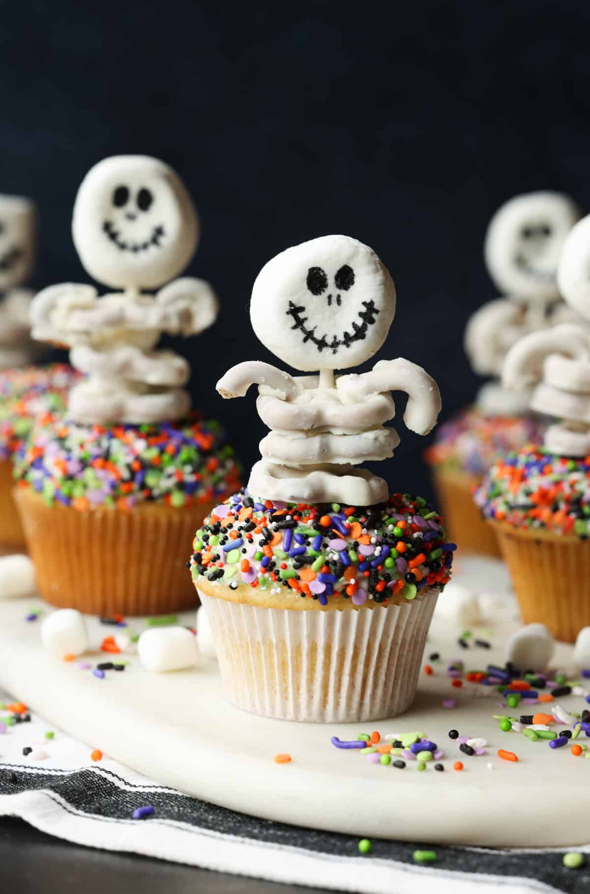 Multiple sprinkle covered cupcakes decorated with skeleton toppers made from white chocolate covered pretzels.