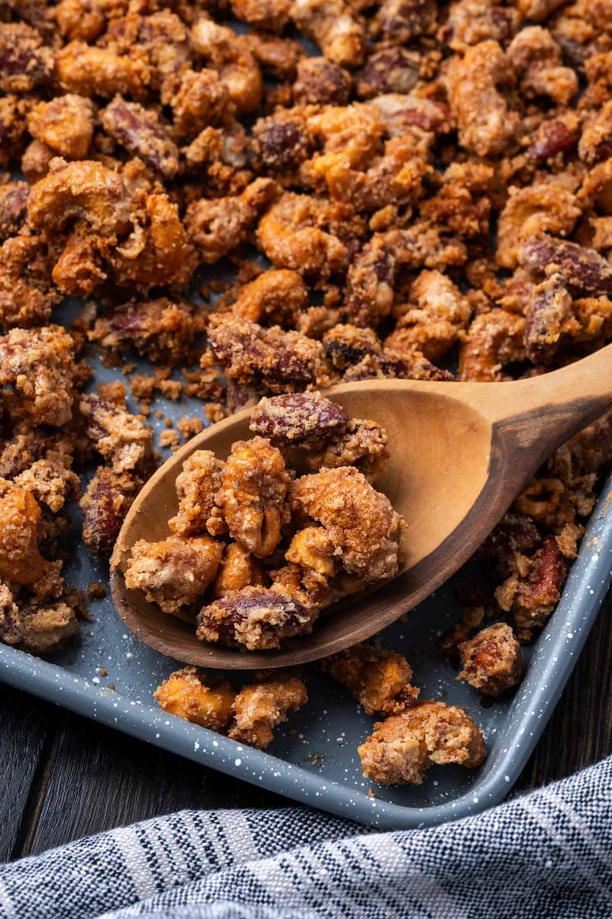 Mashuga nuts are spiced, candied mixed nuts coated in a crispy cinnamon meringue. They make a great homemade gift for friends and neighbors! A wooden spoonful of mashuga nuts resting on a baking sheet.