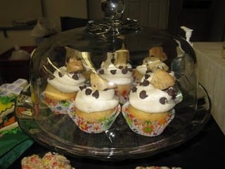 Cookie dough cupcakes on a covered glass cake stand, topped with cookie dough frosting, chocolate chips, and cookie wedges.