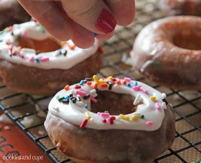 Rainbow sprinkles being added to glazed funfetti donuts on a cooling rack
