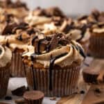 A frosted banana cupcake drizzled with chocolate, with more cupcakes in the background and peanut butter cups scattered in the foreground.