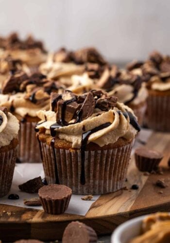 A frosted banana cupcake drizzled with chocolate, with more cupcakes in the background and peanut butter cups scattered in the foreground.