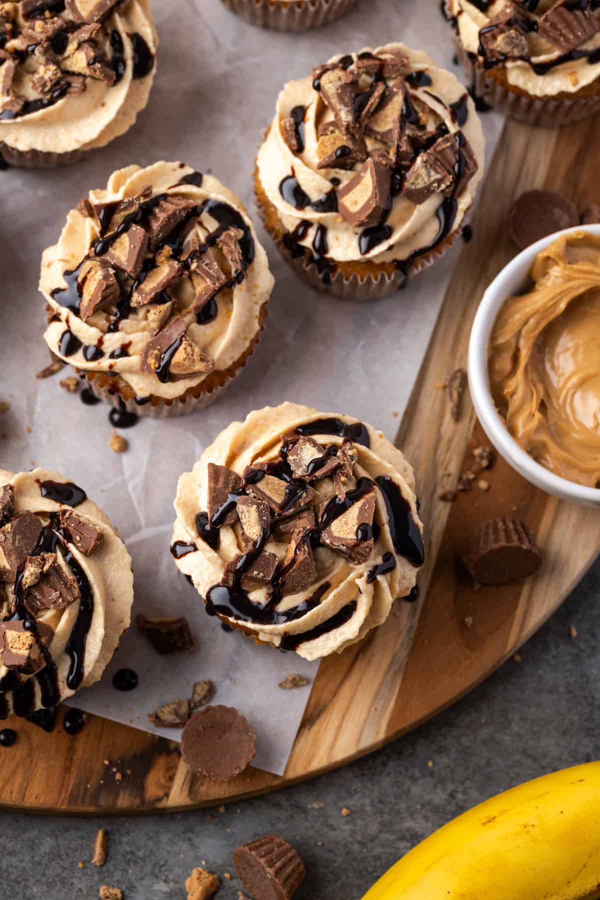 Overhead view of assorted banana cupcakes topped with frosting and a drizzle of chocolate on a parchment-lined wooden board.