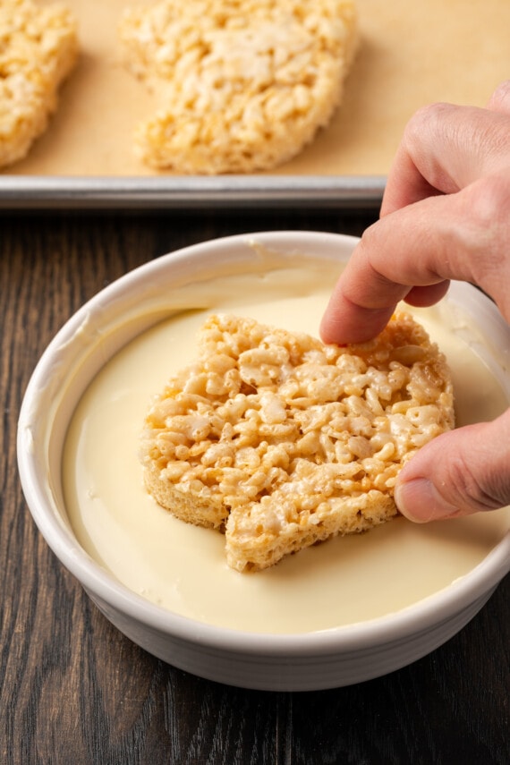 A hand dips a rice krisipie treat ghost into a shallow bowl of melted white chocolate.