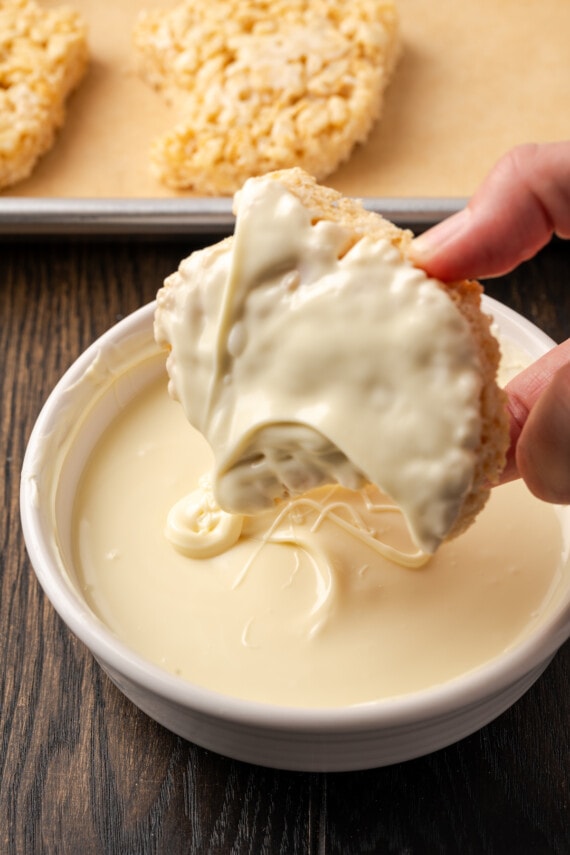 A hand lifts a dipped rice krisipie treat ghost from a shallow bowl of melted white chocolate.