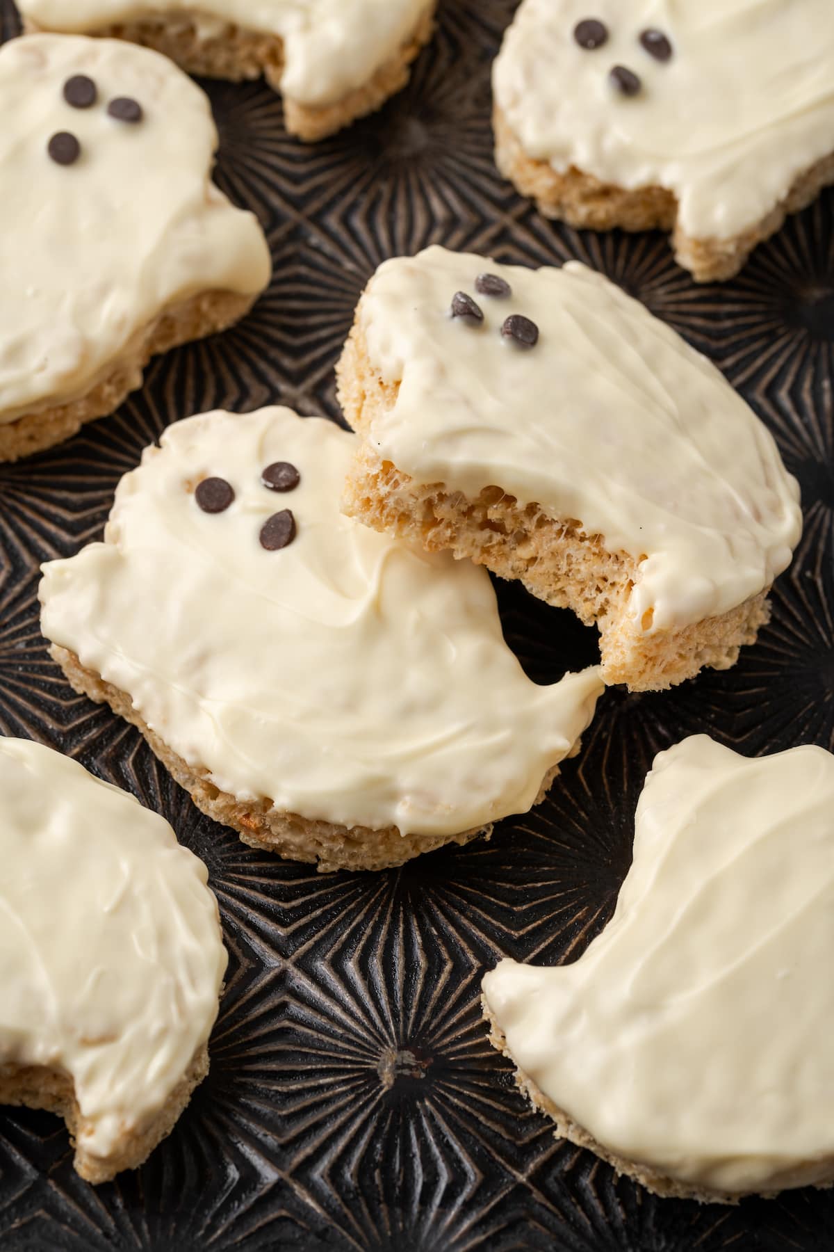 Close up of Halloween rice krispie treat ghosts dipped in white chocolate on a baking sheet, decorated with faces made from mini chocolate chips.