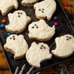 Halloween rice krispie treat ghosts on a baking sheet, decorated with faces made from mini chocolate chips, with a plastic skeleton's fingers in the corner of the frame.