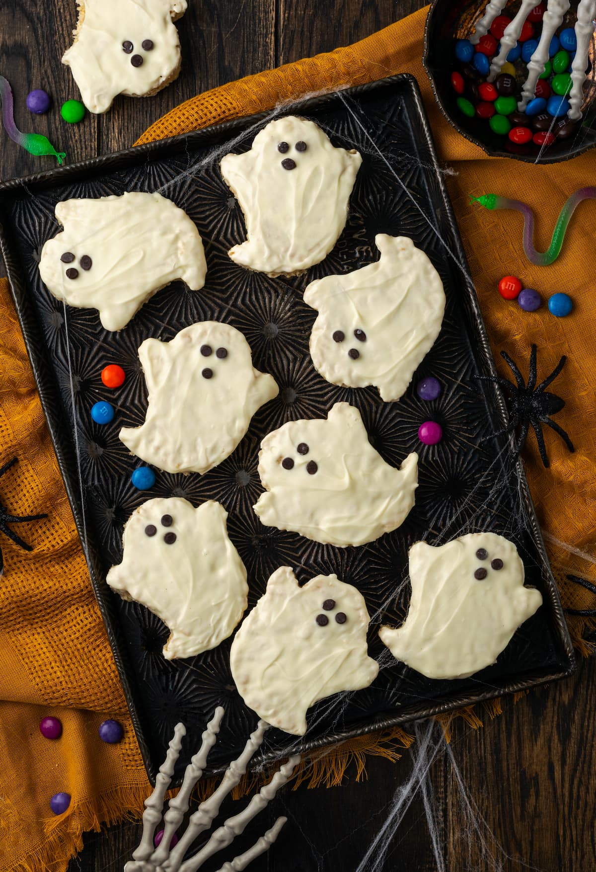 Overhead view of Halloween rice krispie treat ghosts on a baking sheet, decorated with faces made from mini chocolate chips, with a plastic skeleton's fingers in the corner of the frame.