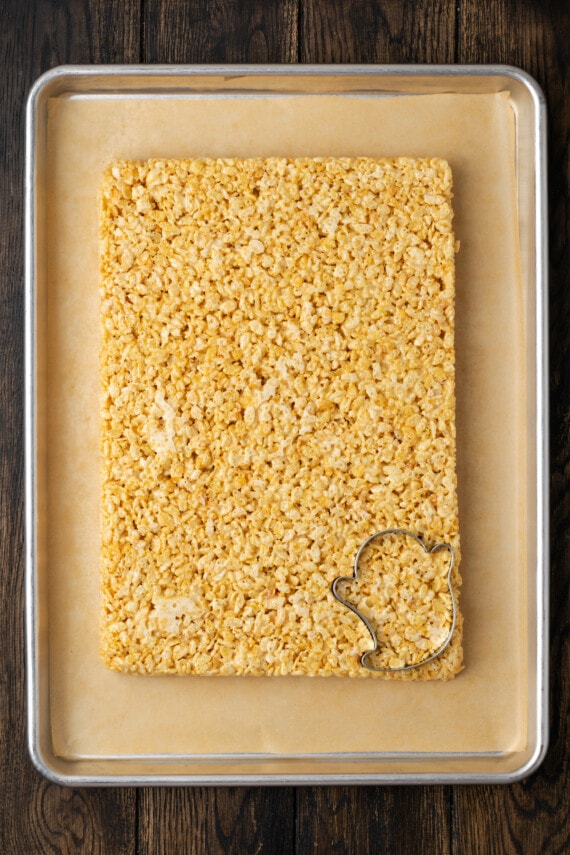 Overhead view of a slab of uncut rice krispie treats on a parchment-lined tray, with a ghost-shaped cookie cutter positioned in the bottom right corner.