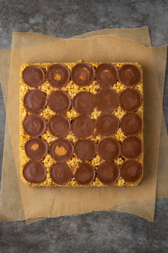 A slab of peanut butter rice krispie treats inverted from the pan onto a piece of parchment paper, with a layer of peanut butter cups on top.