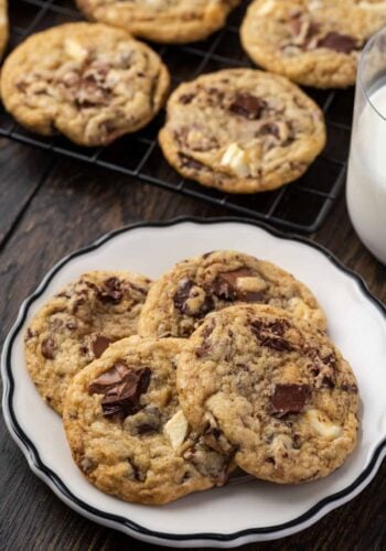 Triple chocolate cookies stacked on a white plate with more cookies on a wire rack next to a glass of milk in the background.