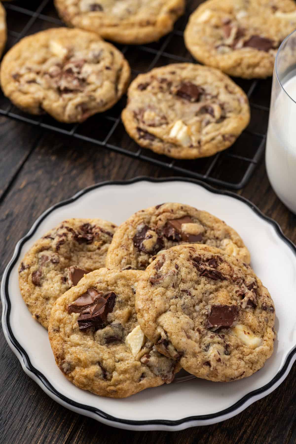 Triple chocolate cookies stacked on a white plate with more cookies on a wire rack next to a glass of milk in the background.