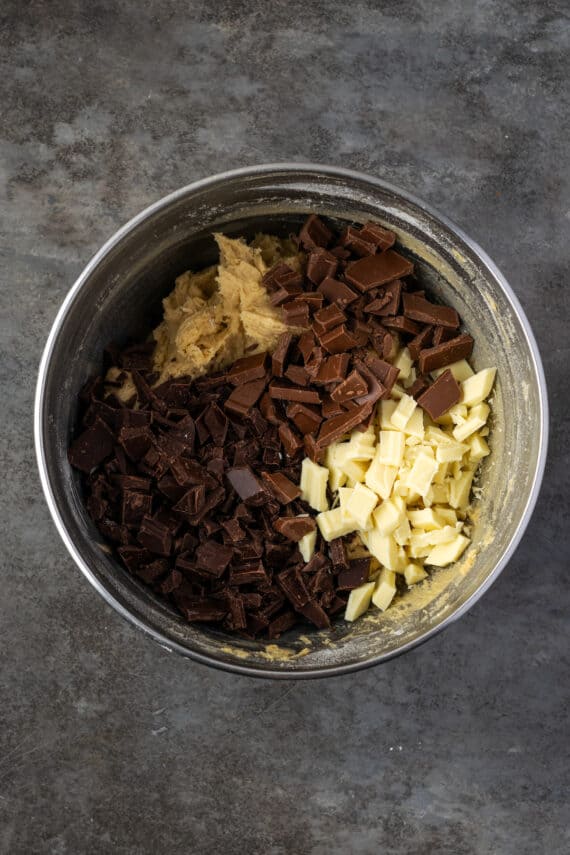 Semi-sweet, milk, and white chocolate chips added to cookie dough in a metal mixing bowl.