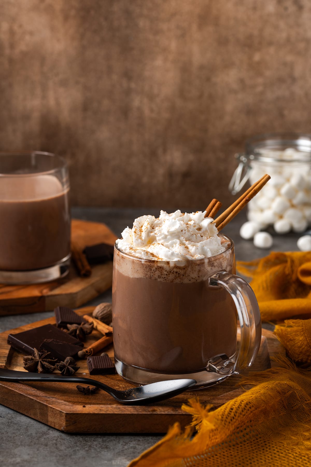Wide shot of a mug of pumpkin spice hot chocolate topped with whipped cream and a cinnamon stick, with a second mug in the background.