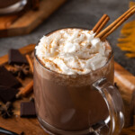 A mug of pumpkin spice hot chocolate topped with whipped cream and garnished with cinnamon sticks.